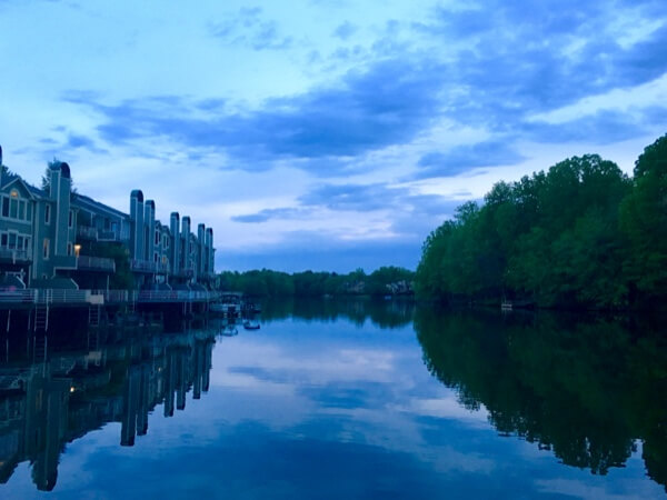 View of a lake meeting the horizon at twilight, with grey panelled houses and trees either side